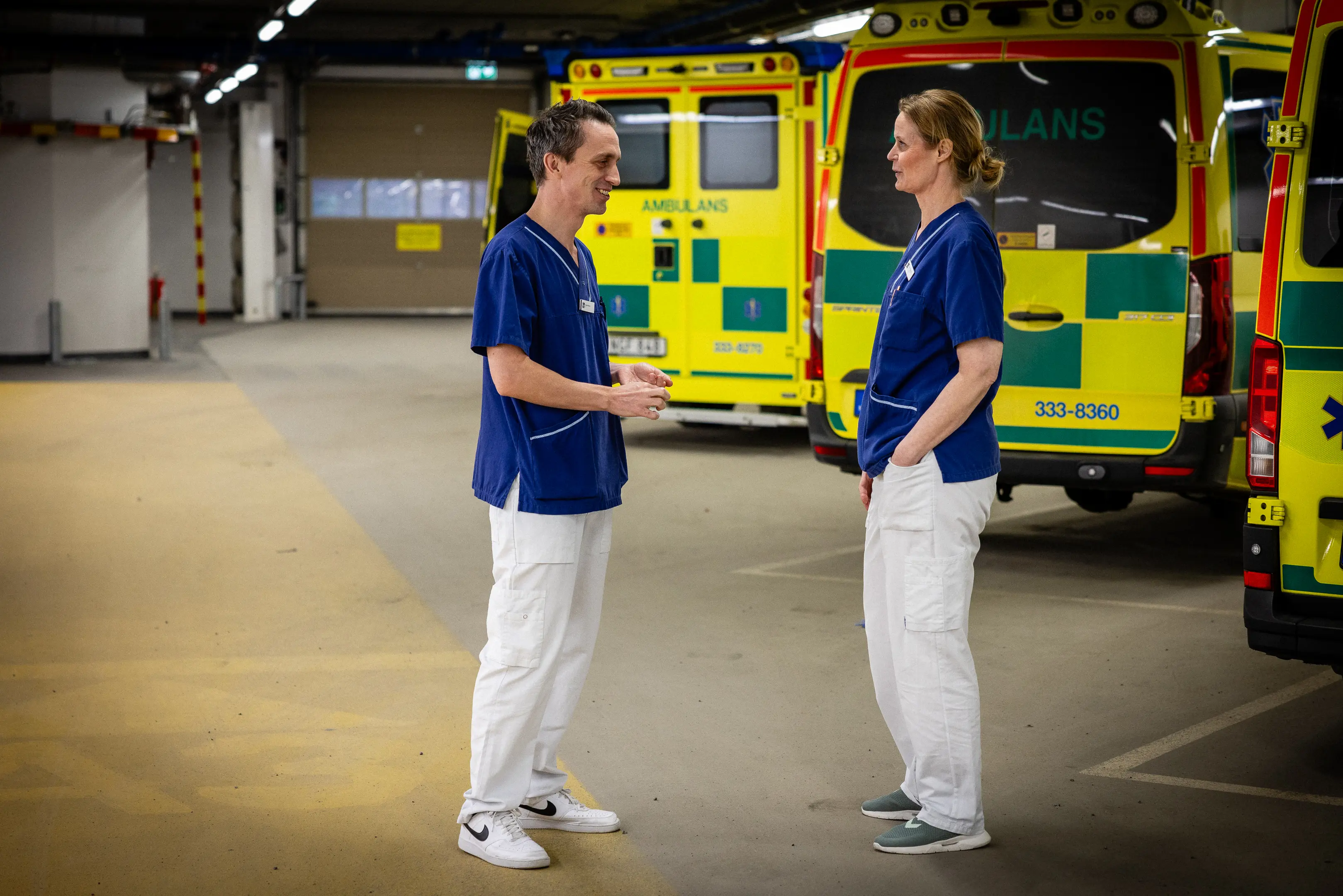 Två medarbetare står i ambulanshallen på sjukhuset framför en rad med ambulanser och pratar. 